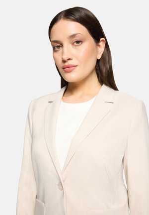 Woman with straight brown hair wearing a cream blazer and white top, facing forward with a neutral expression on a plain background.