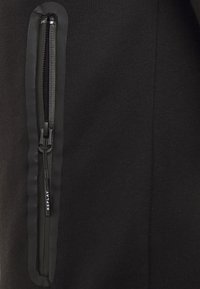 Un tessuto di felpa nera con un piccolo taschino verticale con zip, rifinito con un bordo testurizzato lucido nero e un'etichetta con il logo "REPLAY".