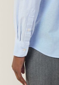 Camisa azul claro con rayas blancas, puños abotonados y una textura suave. Tela gris debajo con sutiles rayas verticales. Sin logotipos ni patrones visibles.