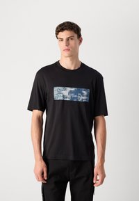 Ung mand iført en sort t-shirt med en blå rektangulær grafisk illustration og hvid "HUGO" tekst, stående foran en ensfarvet lys baggrund.