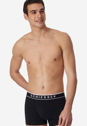Jeune homme souriant, torse nu, portant un boxer noir avec le texte blanc "SCHIESSER" sur la ceinture, debout devant un fond blanc.