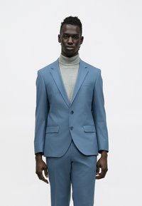 Világoskék öltöny, testhez simuló szabással, két gombbal és oldalzsebekkel, világosszürke garbó fölött viselve. Sima textúra, szabott alak.