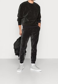 Completo in velluto nero composto da felpa e pantaloni, con polsini e cintura a coste. Abbinato a sneakers grigie. Modello che tiene in mano uno zaino nero.