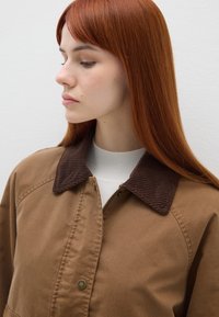 Braune Jacke mit einem gerippten, dunkelbraunen Kragen, ausgestattet mit einem Reißverschluss und Druckknopfverschluss. Der Stoff hat eine glatte Textur und wird über einem weißen Oberteil getragen.