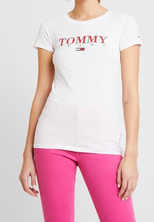 Femme portant un t-shirt blanc Tommy Jeans avec un logo rouge et un pantalon rose vif, debout devant un fond gris clair uni.