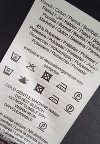 Pflegeetikett auf einem Stoff, mit Pflegeanweisungen, Stoffzusammensetzung (40% Polyester, 60% Baumwolle) und Pflegesymbolen in Schwarz auf weißem Hintergrund.