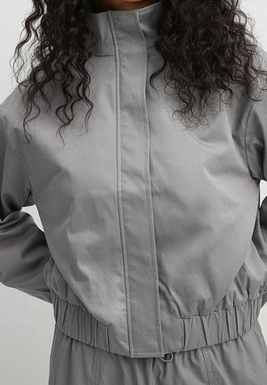 Femme portant une veste gris clair à col montant avec une taille élastiquée froncée, avec de longs cheveux bouclés noirs visibles.