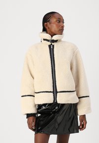 Faux shearling kremfarget jakke med høy krage, svart glidelås og svart trim. Forlommer og brede ermer. Kombinert med et skinnende svart skjørt.
