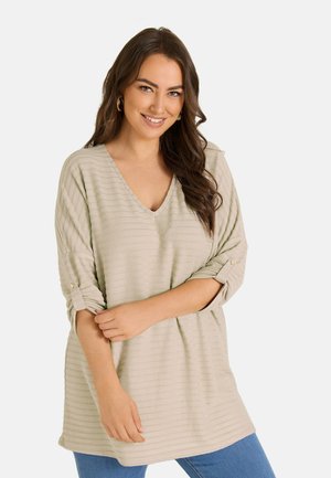 Vrouw met lang bruin haar die een beige gestreepte V-hals top draagt met opgerolde mouwen en een blauwe spijkerbroek, glimlachend met gekruiste armen voor zich.