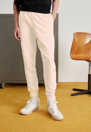 Pantalones deportivos rosa claro con puños y cintura elásticos, con un pequeño logo blanco de Nike. Se llevan con zapatillas blancas de caña alta sobre un suelo amarillo.