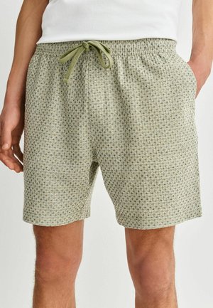 Mann trägt hellgrüne gemusterte Shorts mit Kordelzug und Seitentaschen, Hände an den Seiten, weißes Hemd teilweise sichtbar.