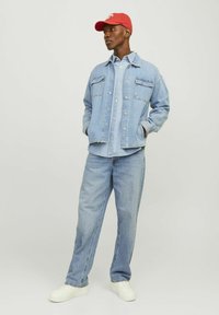  Lichtblauwe denimjack met twee borstzakken, gelaagd over een lichtblauw shirt; gecombineerd met lichtblauwe jeans en witte sneakers; rode pet.