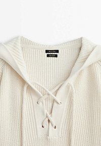 Crème-kleurige gebreide katoen hoodie met een V-halsontwerp, gevlochten voorkant en textiel met ribbelpatronen. Etiket zichtbaar aan de binnenkant.
