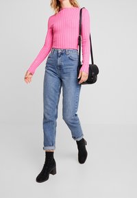 Donna che cammina indossando una maglietta rosa a costine a maniche lunghe, jeans blu a vita alta con risvolti, stivali neri con tacco alla caviglia e una borsa a tracolla nera.