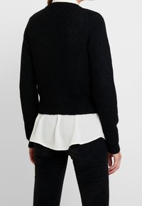 Schwarzer Strickpullover mit gerippter Textur, in einem verkürzten Design. Darunter ist eine weiße, drapierte Bluse zu sehen.