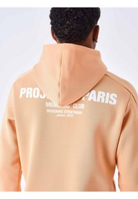 Sudadera melocotón con un gran estampado gráfico blanco en la parte posterior que dice "PROJETS PARIS" y "MEMOIRS' CLUB". Tiene capucha con cordón y una textura suave.