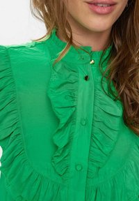 Žalia bluzelė, pasižyminti raukšlelių detalėmis priekyje, su apvaliais mygtukais ir aukštu kaklu. Pagaminta iš lengvos medžiagos, kuri turi švelnų paviršių.