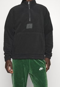 Pullover in pile nero con colletto alto, zip a metà e tasca frontale. Presenta un applicazione testurizzata con un logo, abbinato a pantaloni in velluto verde.