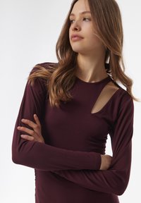 Blusa burdeos de manga larga con escote redondo y corte asimétrico en el hombro izquierdo. Tejido suave con diseño ajustado.