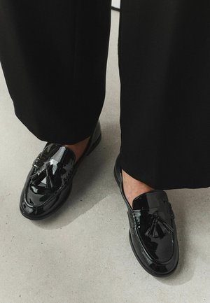 Mocassins à glands en cuir verni noir portés avec un pantalon noir à jambes larges sur un sol gris clair.