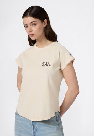 Donna con capelli lunghi e scuri che indossa una maglietta beige a maniche corte con la scritta "S.ATL" e jeans di denim azzurro chiaro, in piedi su uno sfondo bianco.