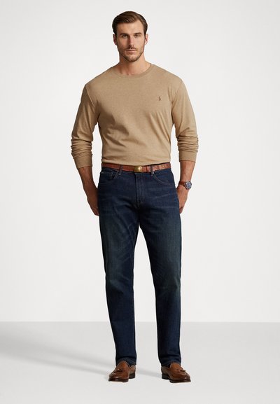 Μπεζ πουκάμισο με μακρύ μανίκι συνδυασμένο με σκούρα μπλε τζιν και καφέ δερμάτινη ζώνη. Το μοντέλο στέκεται σε ουδέτερο φόντο, φορώντας καφέ loafers.