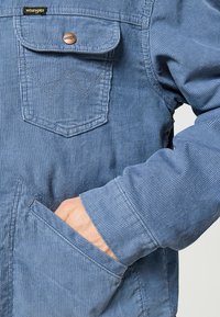 Blå kordjacka med en framficka med varumärkeslogotyp, knappstängning och texturerade ränder på tyget.