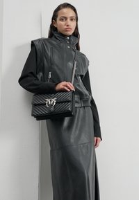 Zwarte leren vest met hoge kraag en ritsen, gecombineerd met een zwarte leren rok. Houdt een gewatteerde zwarte handtas met metalen accenten vast.