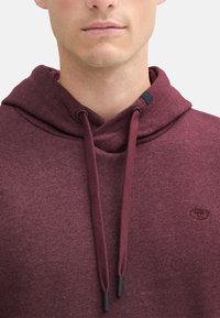 Bordeaux hoodie met een trekkoord, gemaakt van een gestructureerde stof en voorzien van een klein logo op de borst. De capuchon heeft een gelaagd ontwerp voor extra diepte.