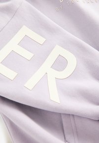 Lilla sweatshirt med store hvide trykte bogstaver. Blødt stof med en glat tekstur og synlige syningsdetaljer.