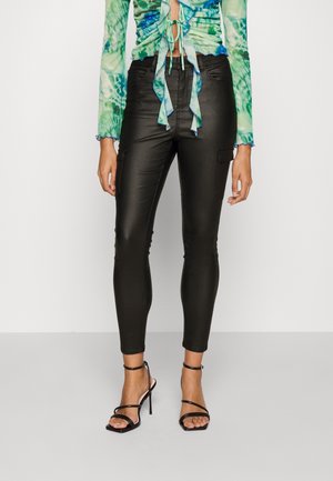 Vrouw die zwarte leren skinny broek, groen-blauw tie-dye geplooide crop top en zwarte sandaal met hoge hakken en bandjes draagt, staand tegen witte achtergrond.
