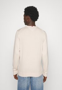 Maglione beige chiaro con orlo e polsini a costine, dotato di una cucitura a forma di V sul retro e di un piccolo dettaglio rosso, bianco e blu al collo.