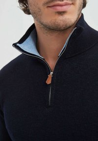 Pull en laine bleu marine avec col zippé. Dessous de col bleu clair ; doté d'une fermeture éclair en métal et d'un tirant en cuir brun.