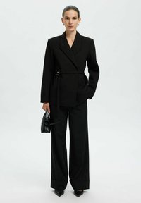 Blazer noir cintré avec une taille ceinturée, associé à un pantalon large noir. Modèle tenant un petit sac à main noir, se tenant devant un fond clair.