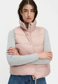 Puffwest in Hellrosa mit hohem Kragen, Reißverschluss vorne und gestepptem Muster. Kombiniert mit einem grauen Langarmoberteil. Angepasste Passform und erkennbare Texturdetails.