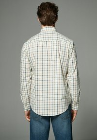 Chemise blanche à manches longues avec un motif à carreaux en vert, orange et bleu marine, dotée d'un col button-down et d'un ourlet arrondi. Portée avec un jean bleu.