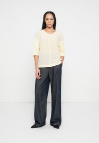 Top galben deschis, semi-transparent, cu mâneci trei sferturi, asortat cu pantaloni largi din denim închis la culoare. Modelul stă cu o mână în buzunar.