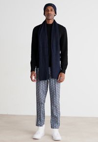 Marineblaue Strickmütze, Schal und schwarzer Pullover kombiniert mit gemusterten blauen Hosen. Weiße Sneakers, mit Fokus auf einheitlicher Textur und Formen.