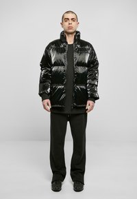 Doudoune noire brillante avec un col montant, design matelassé horizontal et fermeture éclair à l'avant. Portée par-dessus une chemise à manches longues foncée et un pantalon noir.