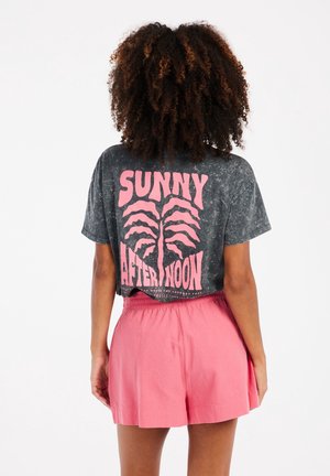 Osoba o kręconych włosach nosi sprany czarny krótki t-shirt z różowym napisem "Sunny Afternoon" i grafiką palmy, połączony z różowymi szortami.