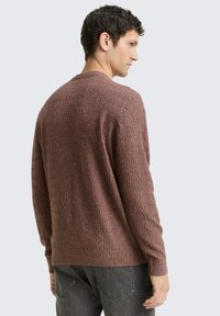 Pull en tricot marron avec un motif texturé, un col rond et des manches longues. Le tissu a un léger brillant et des détails complexes.