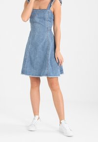 Denim A-linjainen mekko, jossa on neliönmuotoinen pääntie, purjehtivat helmat ja leveät hihnat, yhdistettynä valkoisiin tennareihin rentoon ilmeeseen.