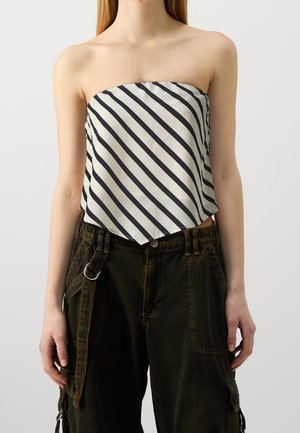 Femme portant un haut sans bretelles rayé en diagonale noir et blanc, associé à un pantalon cargo ample sombre avec une sangle latérale et un anneau en métal.