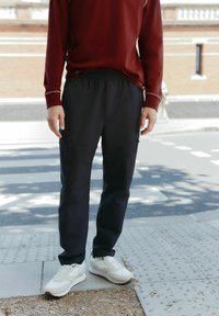 Schwarze Cargo-Hosen mit Reißverschlusstaschen, kombiniert mit einem dunkelroten Langarmshirt. Weiße Sneakers auf einem strukturierten Gehweg.