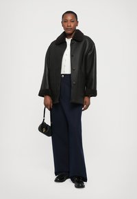 Vrouw in een zwarte schapenvachtjas, witte blouse met knopen, marineblauwe wijde broek, zwarte loafers, met een kleine zwarte handtas in haar hand.