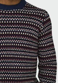 Pull en tricot présentant un design à motifs en marine, bordeaux, noir et blanc. Col roulé et manches longues. Couture texturée sur l'ensemble.