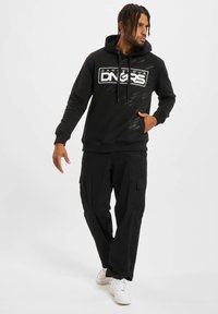 Sudadera negra con estampado de logo, capucha con cordón y bolsillo tipo canguro, combinada con pantalones cargo negros y zapatillas blancas.