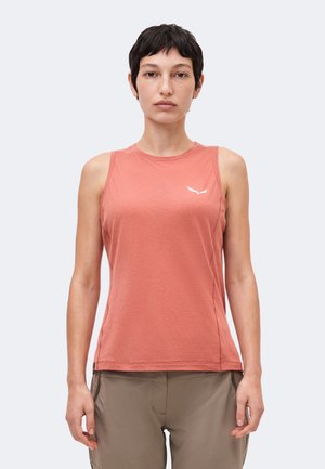 Jeune personne aux cheveux courts et foncés, portant un haut corail délavé sans manches et un pantalon beige, debout devant un fond clair uni.