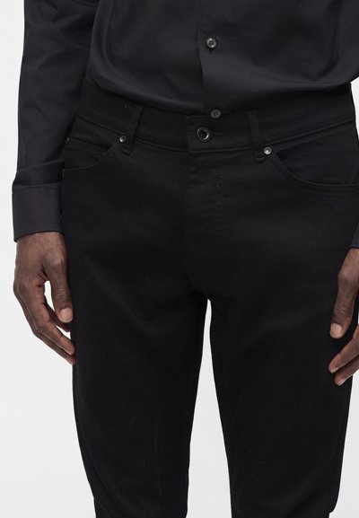 Uomo che indossa jeans neri e camicia nera abbottonata, con le mani rilassate ai lati, mostrato dalla vita a metà coscia.