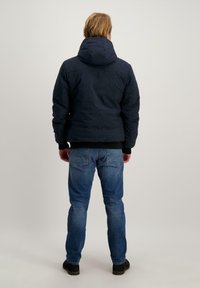 Marineblauwe pufferjack met capuchon, met een gladde buitenkant en ribbels bij de manchetten, gecombineerd met blauwe jeans en zwarte schoenen. Achteraanzicht.